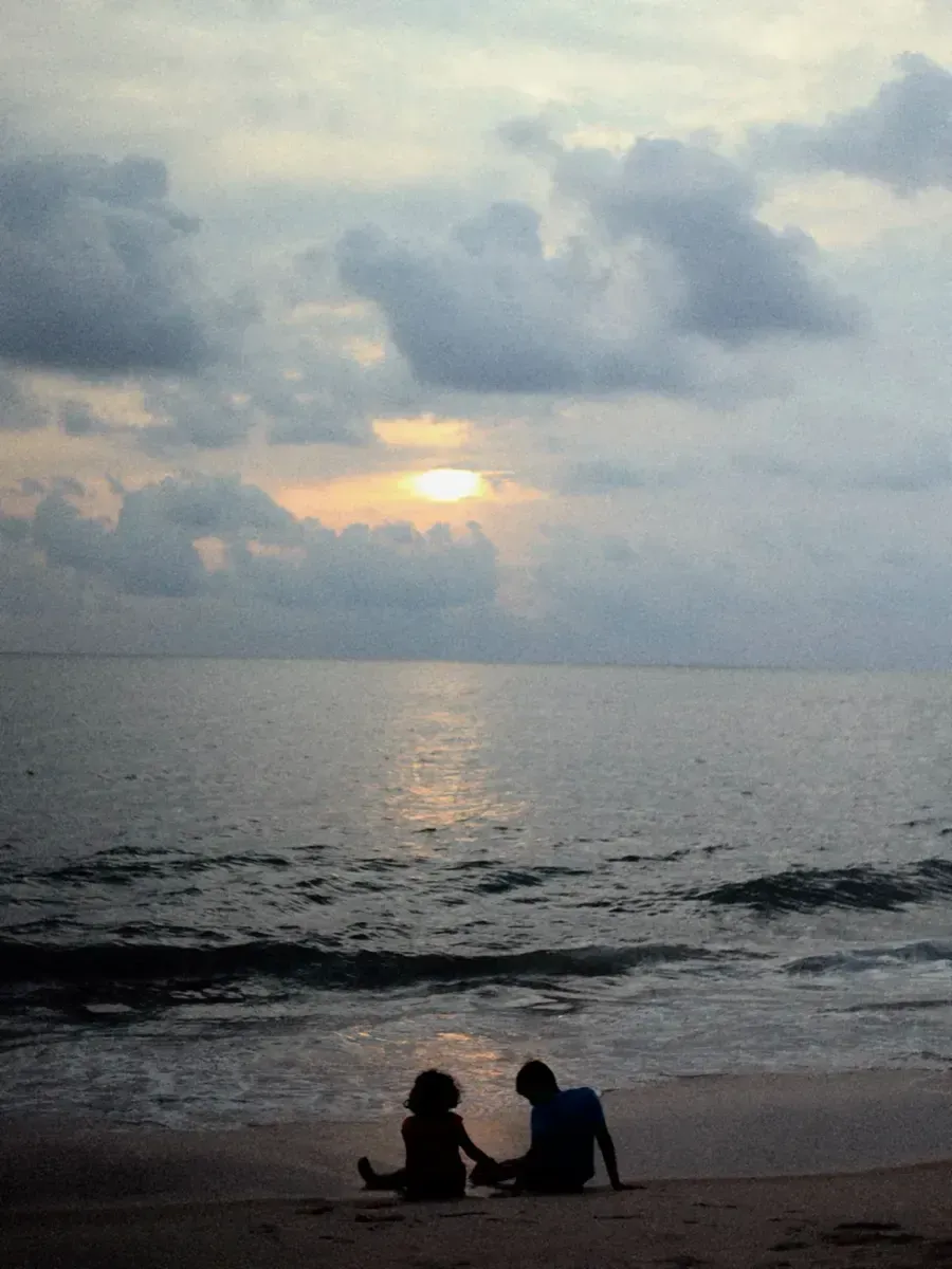 Two people sitting on a beach watching the sunset over the water. Photo by Kiran Johns
