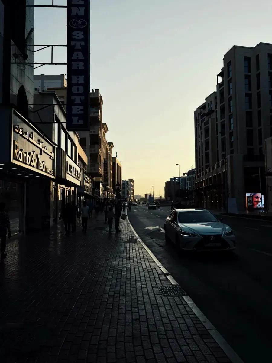 City street at sunset with a car and silhouettes on the sidewalk. Photo by Kiran Johns