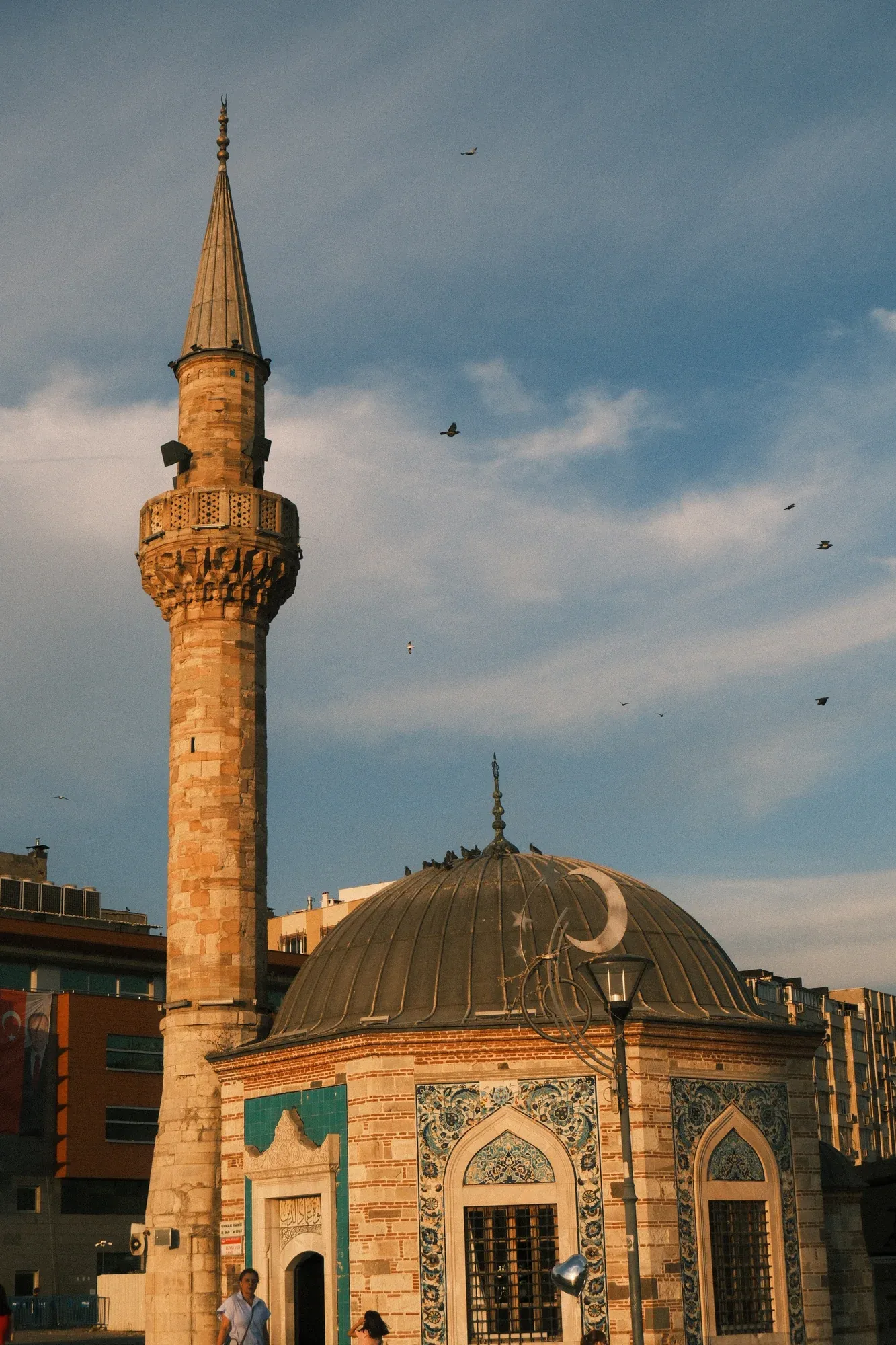 Birds flying above the dome and minaret of a mosque in Konak in golden evening light