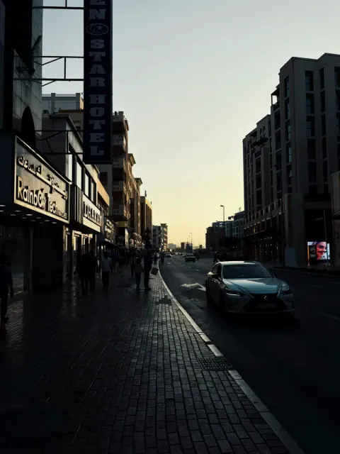 City street at sunset with a car and silhouettes on the sidewalk. Photo by Kiran Johns
