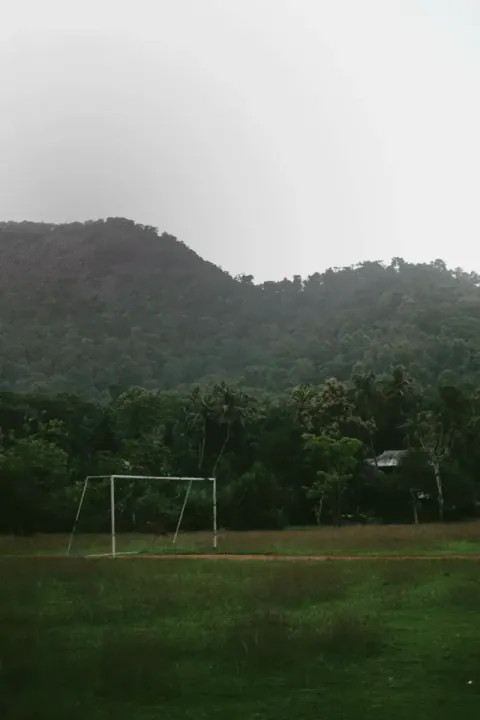White soccer goal on a grassy field with a misty forested hill behind. Photo by Kiran Johns