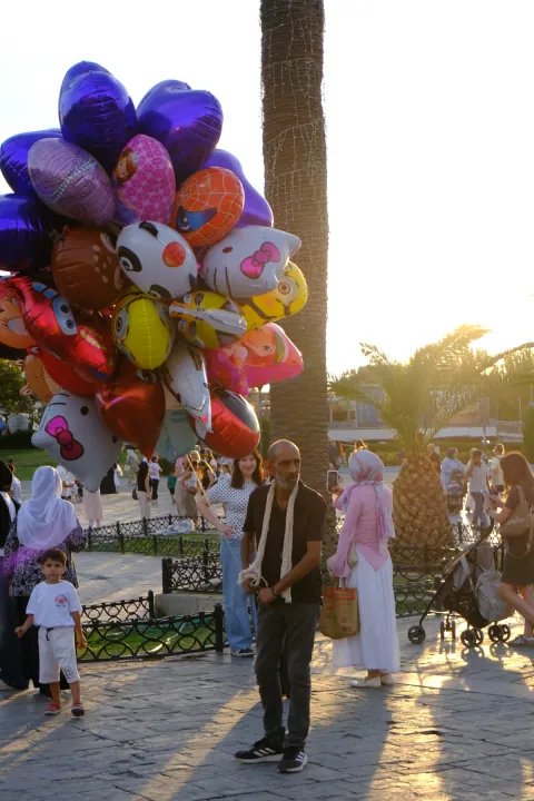A balloon seller holds colourful character balloons in a busy palm-lined square at sunset.