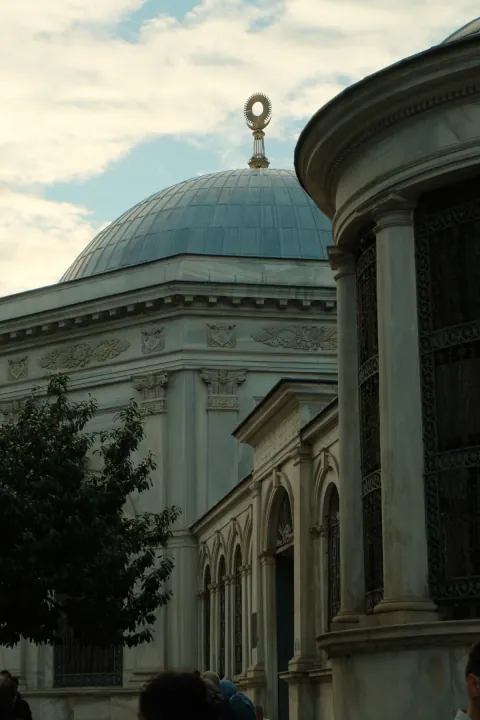 A pale stone dome and ornate columns framed by a curved facade and a cloudy sky.