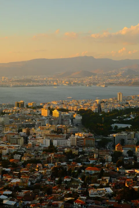 Golden evening light falls across Izmir rooftops, with the bay and mountains stretching into the distance.