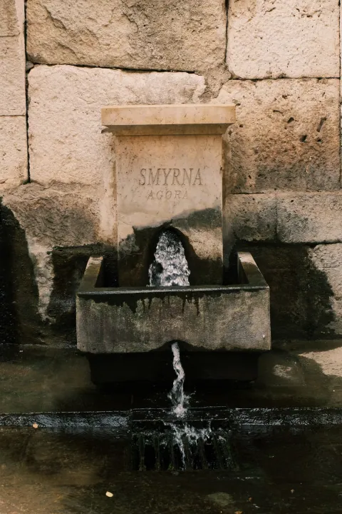 Water pours from an old stone fountain inscribed Smyrna Agora into a weathered basin.