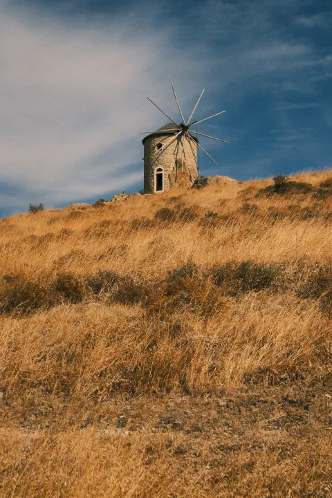 An old round stone windmill with bare wooden spokes stands on a golden grassy hillside beneath a blue sky.